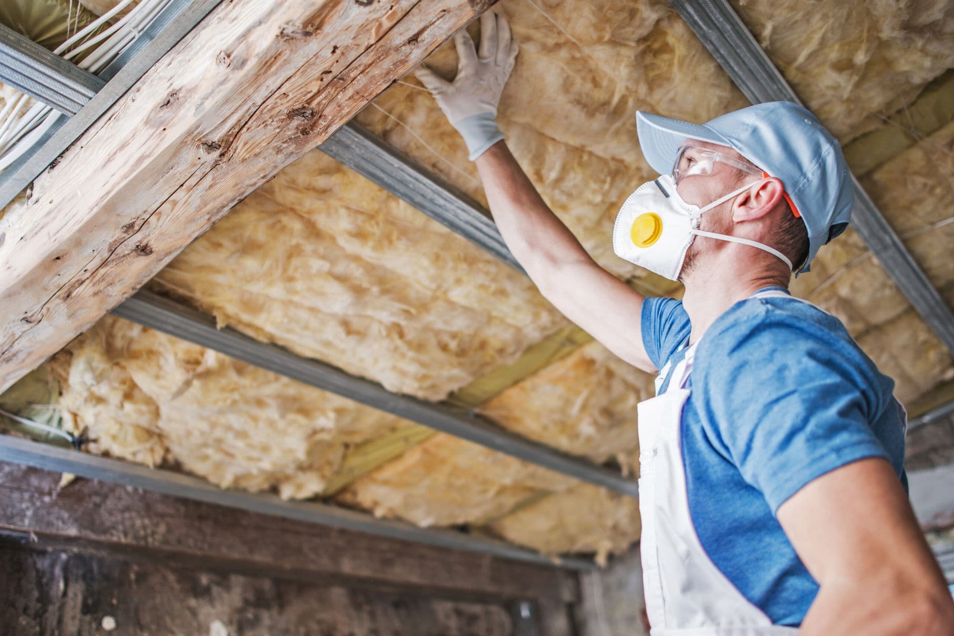 Worker in safety gear installing insulation batts between flooring joists during home construction or renovation.