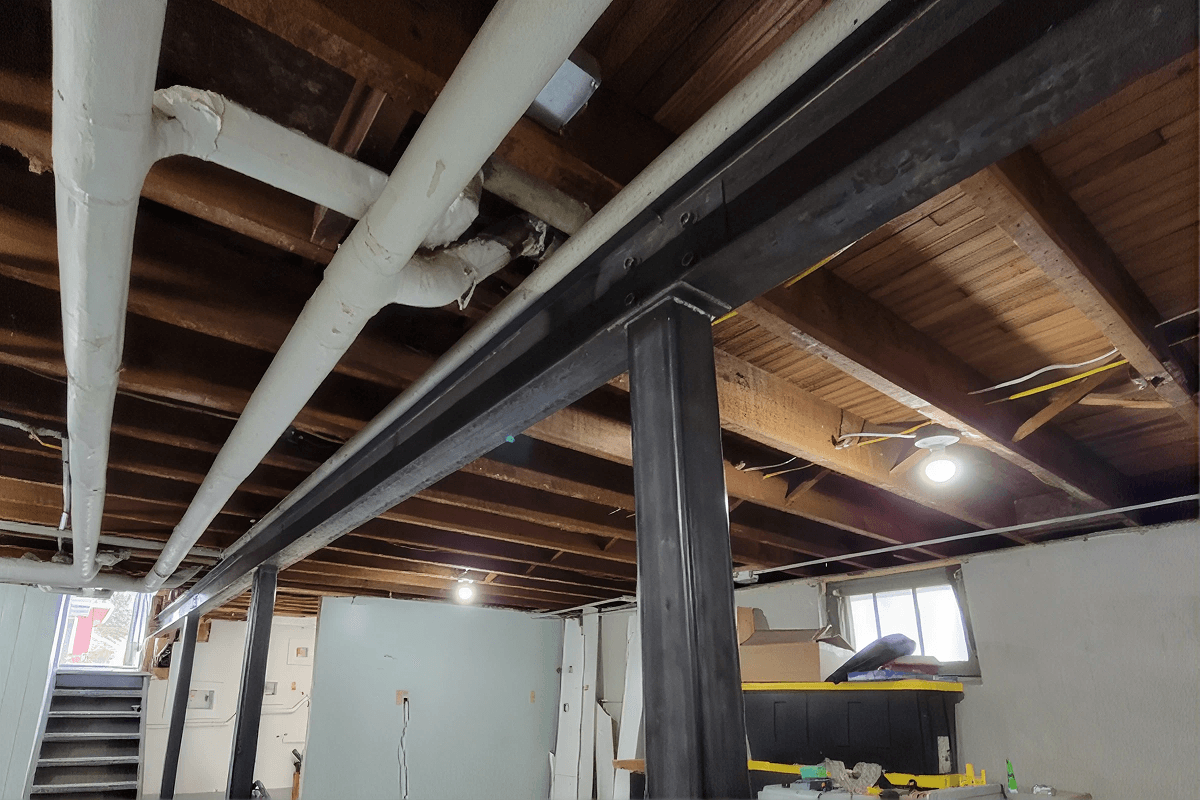 Basement ceiling with exposed wooden joists, white and black beams, recessed lighting, and a new central steel support column.